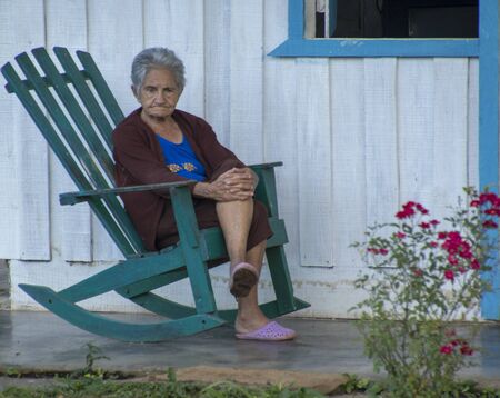 Trinidad, Cuba, Nov 22, 2017 - Old woman sits on green tocking chair on porch against white wallのeditorial素材