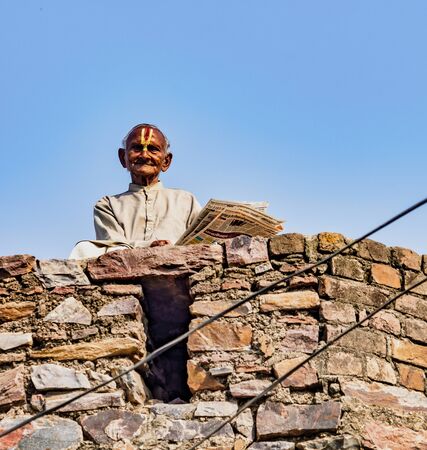 Barsana, India - February 23, 2018 - Old man sits on top of wall reading newspaper and watching people walk byのeditorial素材