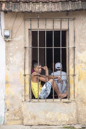 Trinidad, Cuba, Nov 27, 2017 - Boy sits on window sill behind iron bars looked at the streetのeditorial素材