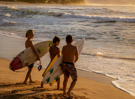 Waikiki, Honolulu, Hawaii - Oct 31, 2021-Young boys hold their surfboards while looking out at the waves at sunset.のeditorial素材