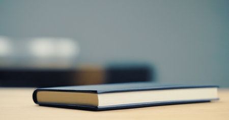 Single, closed, dark blue hardcover book lies on a light wooden surface, with a blurred background.の写真素材