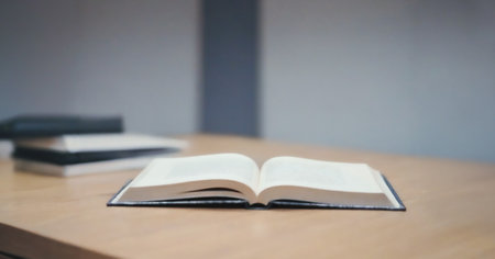 Single open book lies on a light brown wooden table with other books blurred in the background.の写真素材