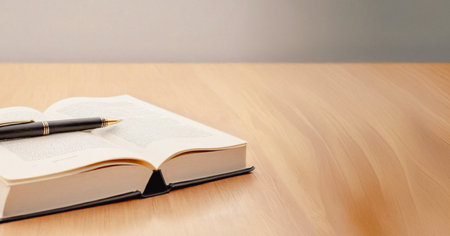 Hardcover book lies open on a light wood surface with a black pen resting on its pages. Premium...