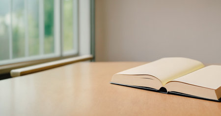 Hardcover book lies open on a light wood table near a blurred window suggesting a quiet space for reading or studying.の写真素材