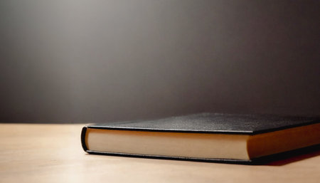 Single, closed, black hardback book sits on a light brown wooden surface against a dark background.