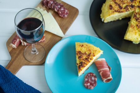 Piece of potato omelette with ham and sausage on a blue plate with a glass of wine on a white background. Copy space.の写真素材