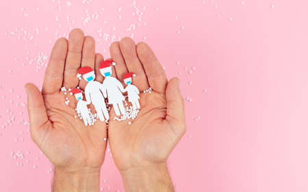 Hands holding a family cut out of paper with a mask and Christmas hat on a pink background.の写真素材