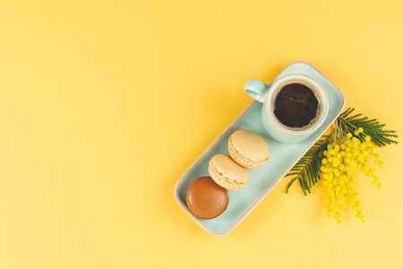Coffee mug with macarons and yellow flowers decoration on yellow background. Copy space. Top view....