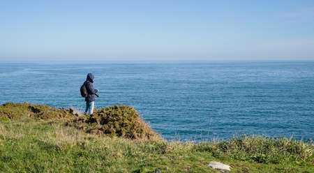 Coastal landscape and man in dark coat with hood and camera looking at calm sea. Copyspace.の写真素材