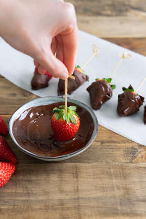 Strawberries with chocolate on a wooden background. Hand dipping strawberry in chocolate bowl. Copy space. Vertical photos.の写真素材