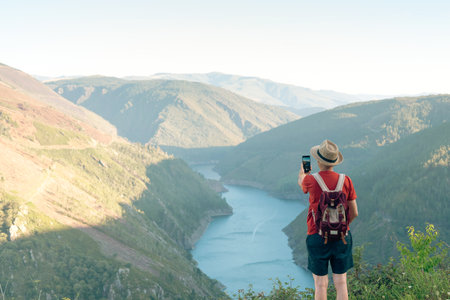 Man with hat and backpack on a sunny day taking photo of landscape with river with cell phone. Copyspace.の写真素材