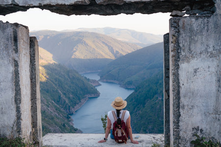 Woman sitting with hat and backpack looking at the landscape of mountains and river on a sunny day from the window of a ruined house. Copyspace. Paicega. weightの写真素材
