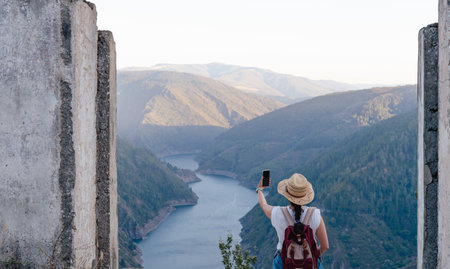 Woman with backpack and hat taking selfie in mountainous landscape. Navia river, Salime reservoir. Paicega. Copyspace. Landscape photo.の写真素材
