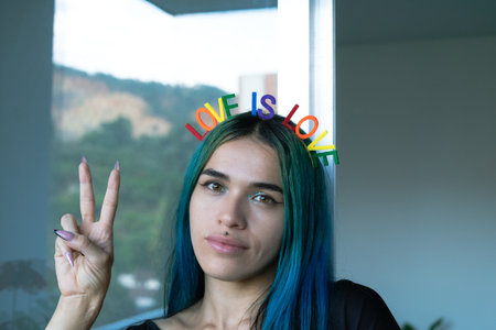 Young woman with serene and pleasant face with blue hair making victory sign with right hand. LOVE IS LOVE headband. Gay theme. Copyspace.の写真素材