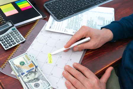A man is writing on a calendar with a pen and a stack of money. The calendar is for the month of April and the man is writing on it.の写真素材