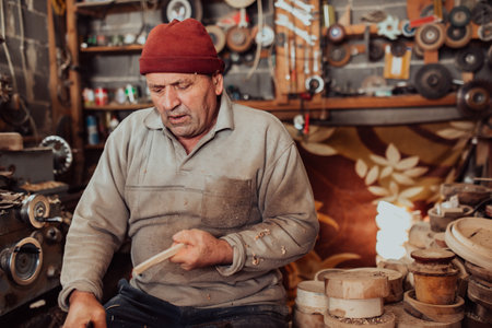 A senior man sitting in the workshop and processing wooden utensils in the old manual way