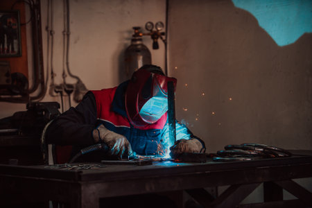 Profesional welder in protective uniform and mask welding metal pipe on the industrial table with other workers behind in the industrial workshop.の写真素材