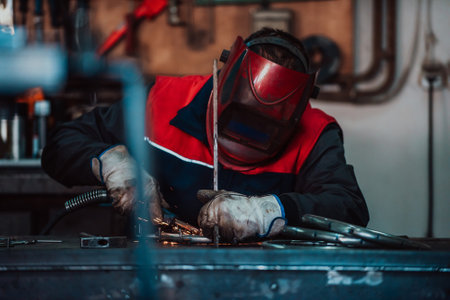 Profesional welder in protective uniform and mask welding metal pipe on the industrial table with other workers behind in the industrial workshop.の写真素材