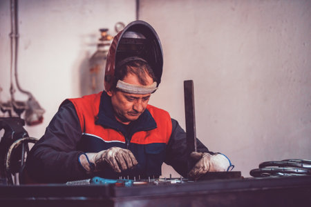 Profesional welder in protective uniform and mask welding metal pipe on the industrial table with other workers behind in the industrial workshop.の写真素材