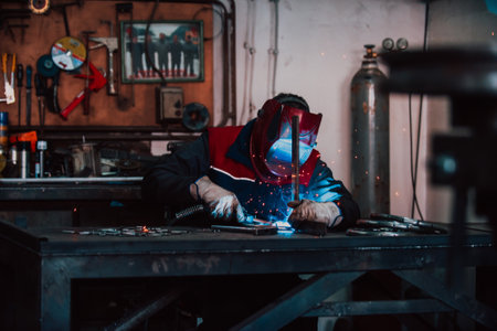 Close up welder in protective uniform and mask welding metal pipe on the industrial table with other...