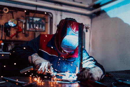 Profesional welder in protective uniform and mask welding metal pipe on the industrial table with...