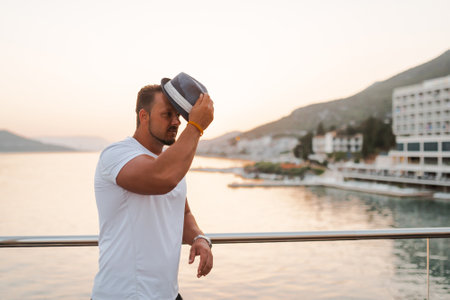 A fit and attractive man on vacation at a hotel near a sandy beach. He is wearing a stylish hat and...