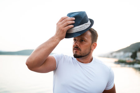 A fit and attractive man on vacation at a hotel near a sandy beach. He is wearing a stylish hat and enjoying the warm sun and crystal-clear sea. The scenery is serene and idyllic, portraying a sense of freedom, simplicity, and pleasure that comes with an active holiday.の写真素材