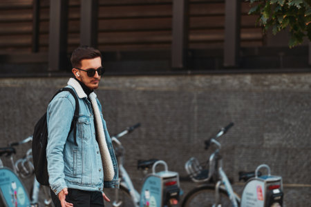 Student walks through the city wearing sunglasses, carrying a backpack, and using wireless headphones, embodying the modern urban lifestyle and the ability to stay connected while on-the-go.の写真素材