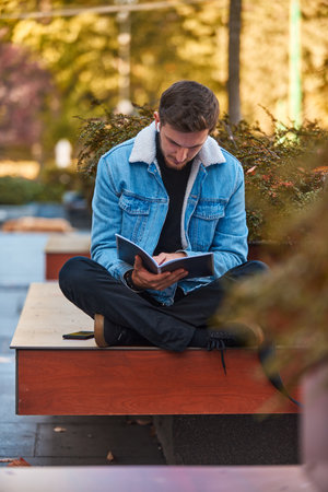 A man sitting on a bench in the city center reading a book while wearing wireless headphones,...