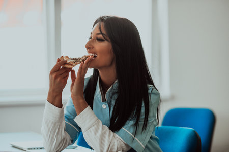 A nurse taking a break from work in the medical office, enjoys a slice of pizza, finding a moment of relaxation and satisfaction amidst her busy healthcare profession.の写真素材