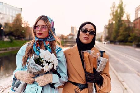 Couple woman one wearing a hijab and a modern yet traditional dress, and the other in a blue dress and scarf, walking together through the city at sunset. One carries a bouquet and bread, while the other holds a blue handbag, symbolizing a beautiful fusion of cultures, friendship, and serene urban charmの写真素材