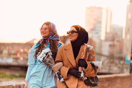 Couple woman one wearing a hijab and a modern yet traditional dress, and the other in a blue dress and scarf, walking together through the city at sunset. One carries a bouquet and bread, while the other holds a blue handbag, symbolizing a beautiful fusion of cultures, friendship, and serene urban charmの写真素材