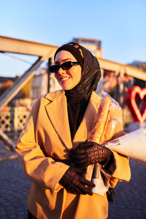 A hijab woman in stylish sunglasses and an elegant French outfit, walking through the city at sunset, carrying a bouquet, bread, and newspaper, radiating a sense of cultural charm and serenityの写真素材