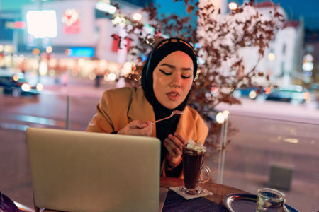 A hijab woman enjoying a sweet dessert at a coffe while working on her laptop during the nighttime in a bustling urban setting.の写真素材
