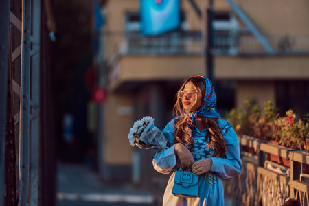 Charm of a woman adorned in an exquisite blue traditional dress, carrying a blue handbag and a bouquet of flowers, gracefully strolling through the city at sunset, creating a mesmerizing scene of elegance, cultural richness, and the tranquil ambiance of dusk.の写真素材
