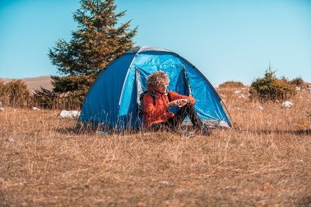 A man camping with a tent. He is surrounded by a beautiful mountain landscape, and the sun shines on his face, foreshadowing a beautiful and sunny day. The man looks satisfied and fulfilled, suggesting that he is enjoying his adventurous journey.の写真素材