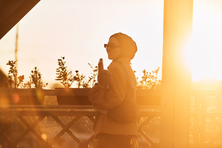 A hijab woman in stylish sunglasses and an elegant French outfit, walking through the city at sunset, carrying a bouquet, bread, and newspaper, radiating a sense of cultural charm and serenityの写真素材