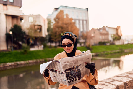 A woman in a hijab gracefully adorned in a fusion of old and modern French attire, immersed in the bustling streets of the city, engrossed in reading a newspaperの写真素材