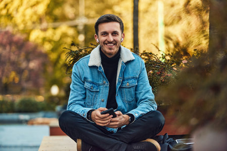 A man sitting on a bench in the city center using his smartphone and wireless headphones for a call, showcasing the convenience of technology in modern communication and the ability to multitask while on-the-go.の写真素材