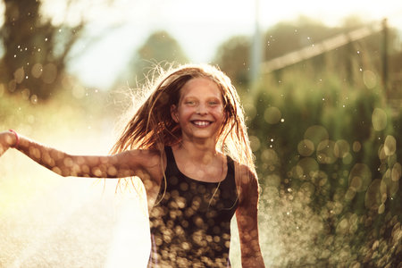 In the backyard of house, a young girl bursts with laughter and joy as she gleefully runs through a water sprinkler on a sunny summer dayの写真素材