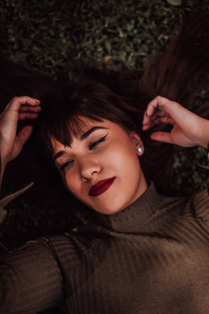 A young woman is lying on the grass, showing off her beautiful face with a serene expression. The lush greenery in the background adds to the peaceful atmosphere of the scene, making it a relaxing and idyllic moment captured in time.の写真素材