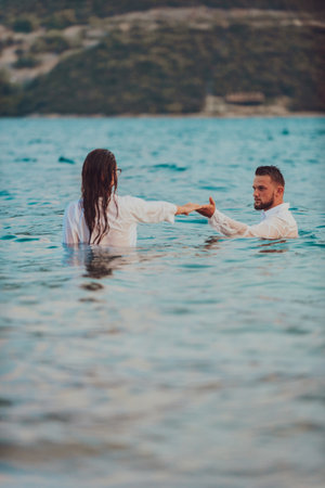 A young couple, dressed in attire, symbolically holds hands while standing in the sea during the mesmerizing sunset, creating a picturesque scene of love and unity against the backdrop of the ocean horizonの写真素材