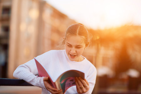 A beautiful blonde teenager sits on a bench in the park, taking a break from school to immerse herself in a book, accompanied by her school backpackの写真素材