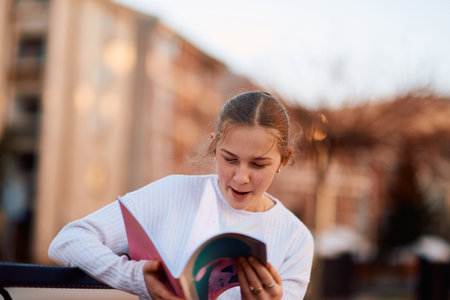 A beautiful blonde teenager sits on a bench in the park, taking a break from school to immerse herself in a book, accompanied by her school backpackの写真素材