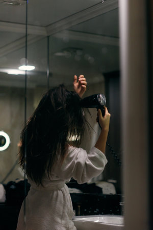 A joyful woman, with headphones in her ears, playfully dries her hair with a hairdryer in the bathroom after a refreshing bath, embodying a carefree and energetic moment of self-care and relaxationの写真素材