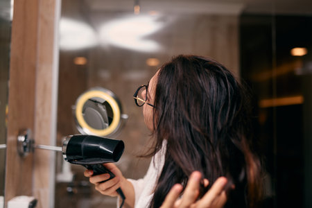 A joyful woman, with headphones in her ears, playfully dries her hair with a hairdryer in the bathroom after a refreshing bath, embodying a carefree and energetic moment of self-care and relaxationの写真素材