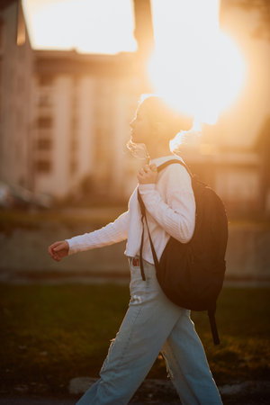 A beautiful European teenager, captured in a side-profile portrait, walks home during sunset after school, carrying her backpackの写真素材