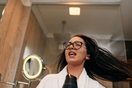 A joyful woman, with headphones in her ears, playfully dries her hair with a hairdryer in the...
