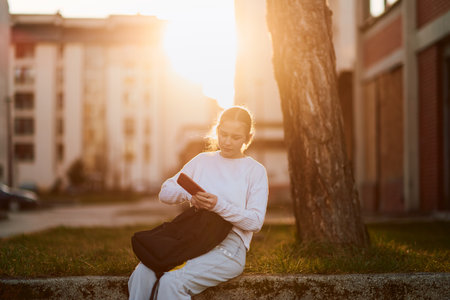 A beautiful blonde teenage girl sits on a park bench, using her smartphone and wireless headphones to prepare for school, with her school backpack beside her on the benchの写真素材