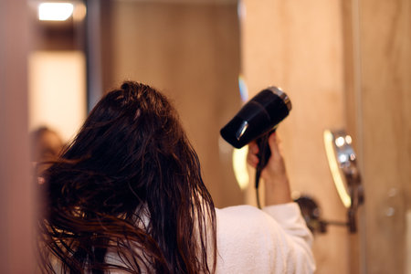 A joyful woman, with headphones in her ears, playfully dries her hair with a hairdryer in the bathroom after a refreshing bath, embodying a carefree and energetic moment of self-care and relaxationの写真素材